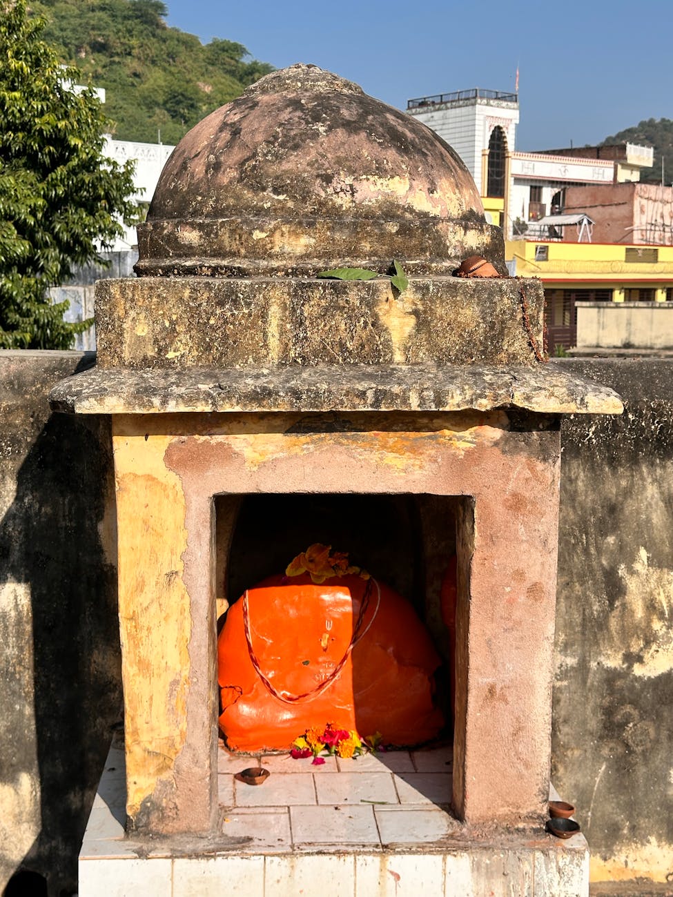 A small outdoor shrine housing a vibrant Hanuman idol in bright sunlight.